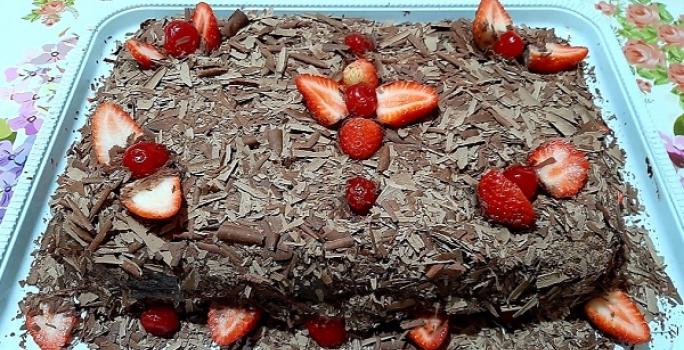 Foto em tons de marrom da receita de bolo de chocolate com morangos servida inteira e decorada com morangos e cerejas em cima