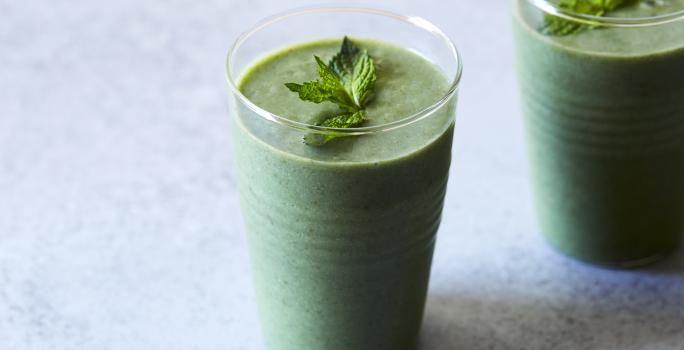 Fotografia de dois copos de vidro com um refresco de hortelã, e por cima, folhas de hortelã. As vitaminas estão sobre uma mesa em tons de azul claro.