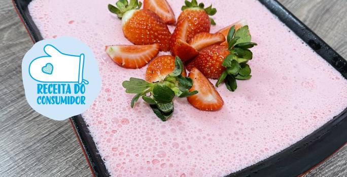 Imagem de uma receita de gelatina em tom rosa, decorada com morangos picados, servida em um refratário preto. Na imagem há um selo com o texto Receita do Consumidor em tom azul claro.