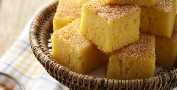 Foto de pedaços quadrados de bolo de fubá em um recipiente marrom em cima de um pano branco com detalhes laranja e azul