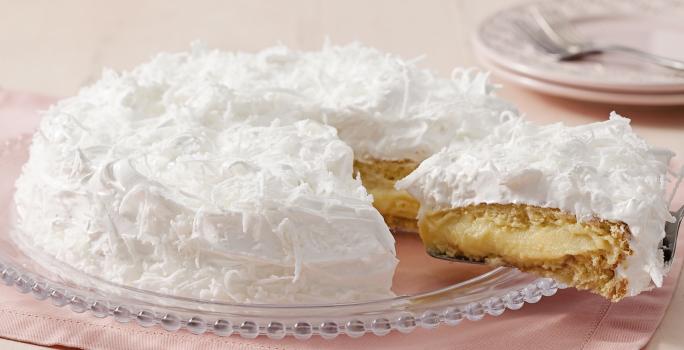 Fotografia de um bolo coberto com marshmallow branco e raspada de coco em cima. Dentro do bolo creme e massa brancos. O prato está em cima de um guardanapo de papel rosa claro.
