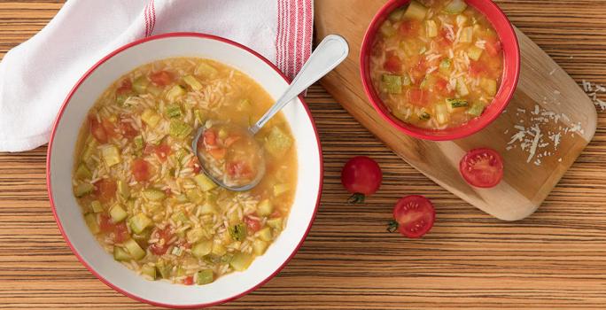 Fotografia em tons de branco, vermelho e marrom, sopeira branca com borda vermelha contendo sopa com legumes, arroz e uma concha, ladeada por guardanapo branco e vermelho, ao lado pote com sopa sobre tábua, tomate, tudo sobre bancada em tons de marrom.