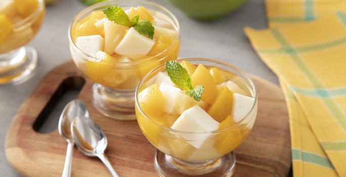 Foto da receita de Gelatina de Verão. Observa-se três potes individuais de vidro com as gelatina em quadrados e frutas amarelas picadas sobre uma tábua de madeira. Ao lado dos potes, duas colherinhas acompanham.