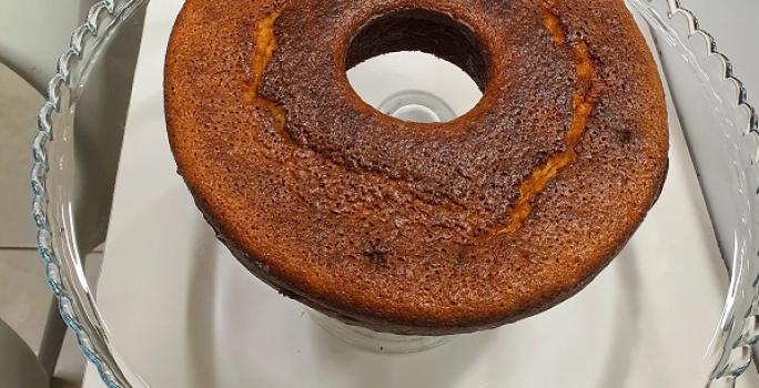 Foto da receita de bolo de cuscuz servida em uma porção grande sobre uma bailarina de vidro em cima de uma mesa branca