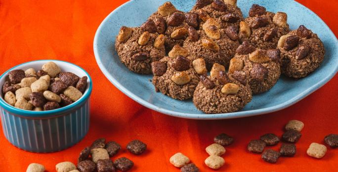 Fotografia em tons de vermelho em uma bancada de madeira de cor vermelha. Ao centro, um prato redondo azul contendo os cookies. Ao lado, há um recipiente contendo passatempo cereal e ao redor há alguns cereais espalhados.