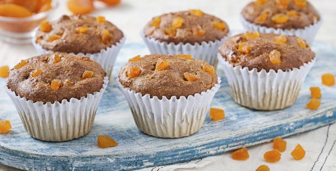 Fotografia em tons de azul em uma bancada de madeira clara, um paninho branco com listras azuis escuras, uma tábua de madeira azul e vários muffins integrais com damasco em forminhas brancas.