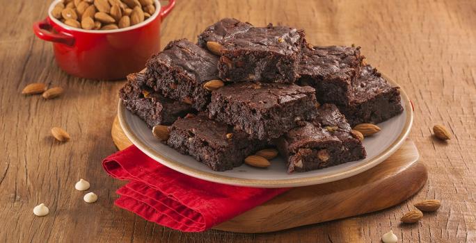 Fotografia em tons de marrom e vermelho de uma bancada de madeira com uma tábua de madeira e um paninho vemelho, sobre ele um prato redondo com pedaços de brownie. Ao fundo um recipiente redondo com amêndoas.