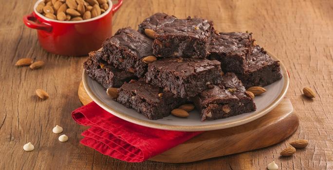 Fotografia em tons de marrom e vermelho de uma bancada de madeira com uma tábua de madeira e um paninho vemelho, sobre ele um prato redondo com pedaços de brownie. Ao fundo um recipiente redondo com amêndoas.