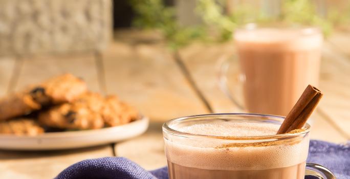 Fotografia em tons de marrom em uma bancada de madeira com um pano azul e duas xícaras de vidro com o chocaccino e um pau de canela para enfeitar. Ao fundo, um prato com cookies