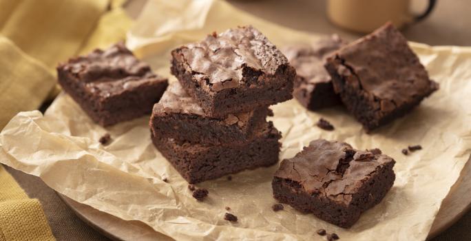 Fotografia em tons de marrom e bege em uma bancada de madeira, um pano bege, uma tábua de madeira redonda, um papel manteiga marrom com vários brownies de chocolate em cima. Ao fundo, xícaras beges.