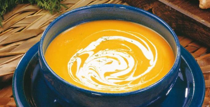 fotografia em tons de laranja, azul e marrom de uma bancada marrom vista de cima, contém um prato redondo azul com um recipiente redondo e azul com a sopa cremosa de cor laranja.