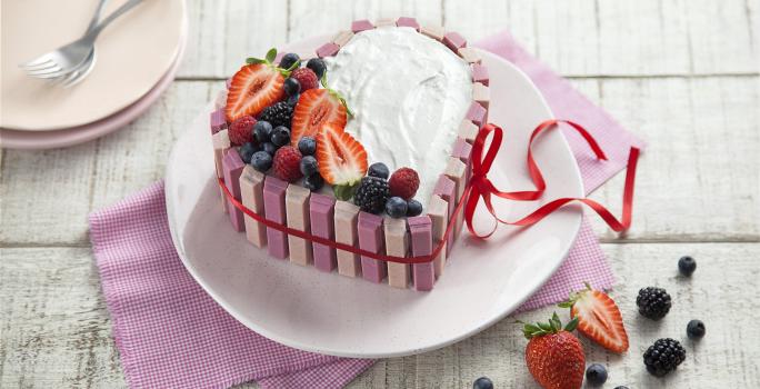 fotografia em tons de branco e rosa de uma bancada branca vista de cima, ao centro um pano rosa com um prato redondo por cima um bolo em formato de coração com marshmallow, morangos e frutas vermelhas por cima para decorar.