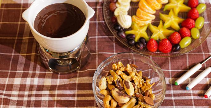 Fotografia em tons de marrom em uma bancada com uma toalha xadrez em marrom, com uma pequena panela branca com o fondue de chocolate dentro, ao fundo um recipiente com frutas picadas e um potinho de vidro com castanhas e amêndoas.