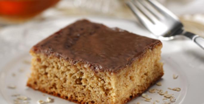 Fotografia em tons de marrom em uma mesa com uma toalha branca e bege, um prato branco raso redondo com uma fatia do bolo de banana com aveia e mel. Ao fundo, um potinho de mel.