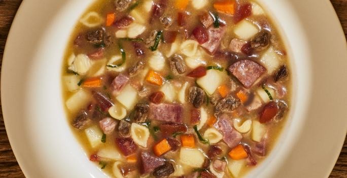 Foto bem aproximada de um prato branco fundo no qual está a receita pronta de sopão de carne com legumes e linguiça