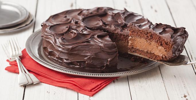 Fotografia em tons de branco e marrom de uma bancada de madeira, um paninho vermelho, um prato redondo prateado, dois garfos prateados e um bolo de chocolate com uma fatia sendo retirada, mostrando o recheio de mousse.