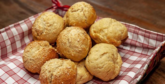 Fotografia de pãezinhos com massa de requeijão e queijo parmesão. Os pães estão dentro de uma cestinha quadrada com papel quadriculado vermelho e branco, que está sobre uma mesa de madeira.