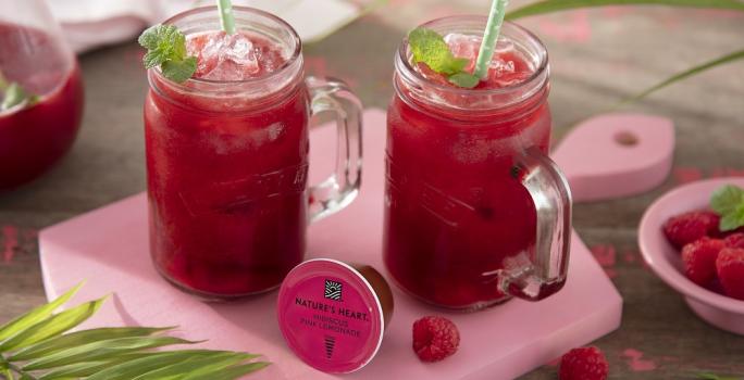 Foto em tons de cor de rosa da receita de pink lemonade com frutas vermelhas e hortelã servida em canecas altas de vidro em cima de uma tábua rosa clarinha com um pote de framboesas ao lado