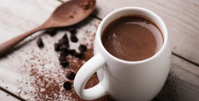 Fotografia em tons de branco e marrom tirada de uma bancada marrom, contém uma xicara branca com chocolate quente dentro e ao lado uma colher de madeira e chocolate em pó espalhado pela bancada.