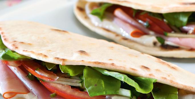 Fotografia em tons de rosa em uma mesa de madeira com um pano rosa florido, uma tábua de madeira branca ao centro com a piadina, que é uma massa estilo crepe francês, recheada de tomate, rúcula e peito de peru.