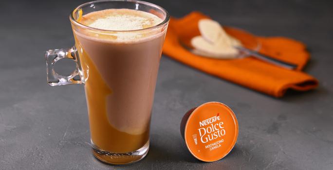 Fotografia em tons de laranja em uma bancada de madeira cinza escura, um paninho laranja e uma xícara de vidro alta com o café de mochaccino canela com doce de leite e achocolatado Galak.