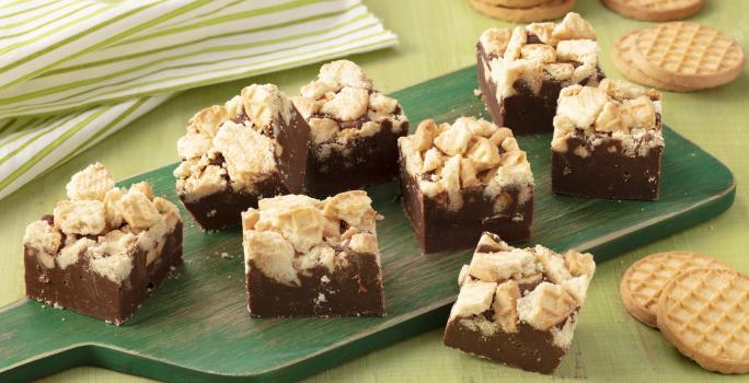 Fotografia em tons de verde de uma bancada verde com uma tábua verde, sobre ela pedaços de fudge. Ao fundo um paninho listrado verde e branco e biscoitos tostines. A frente biscoitos tostines.