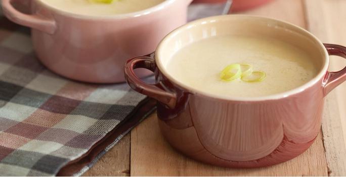 Fotografia em tons de rosa em uma bancada de madeira com uma toalha xadrez preta e rosa, duas mini panelinhas de cerâmica rosa com a sopa de alho-poró dentro delas.