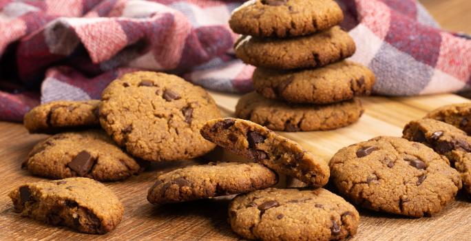 fotografia em tons de marrom, vinho e roxo de uma bancada marrom vista de frente, contém diversos biscoitos de chocolate espalhados pela mesa, e ao fundo  um pano xadrez em tons de vinho e roxo.