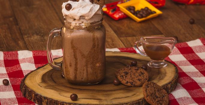 Foto de uma bancada de madeira. Sobre ela há uma toalha quadriculada vermelho e branco, uma tábua de madeira com um copo com o Milkshake de Nescau decorado com um canudo vermelho. Ao lado há um potinho com calda, 2 cookies e algunmas bolinhas de chocolate