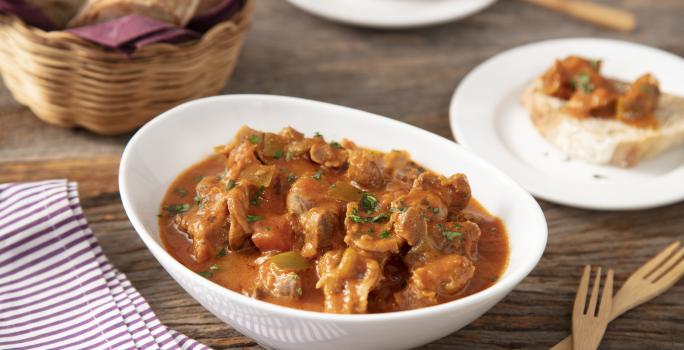 Fotografia em tons de marrom em uma bancada de madeira escura, um paninho roxo listrado, um recipiente branco com a moela de frango com molho de tomate dentro. Ao lado, um cesto de vime com pães, talheres de madeira e um prato menor com uma fatia de pão.