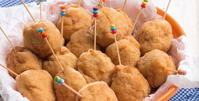 Fotografia em tons de azul em uma bancada de madeira clara com um pano azul quadriculado e em cima um prato redondo fundo laranja com coxinhas cremosas de frango dentro dele e palitinhos coloridos espetados nelas.