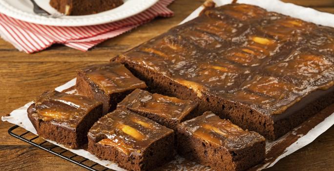 fotografia em tons de marrom e branco de uma bancada marrom vista de frente. Contém uma grade com um papel branco por cima, que contém um bolo de chocolate com pedaços de maçãs e cortado em quadrados