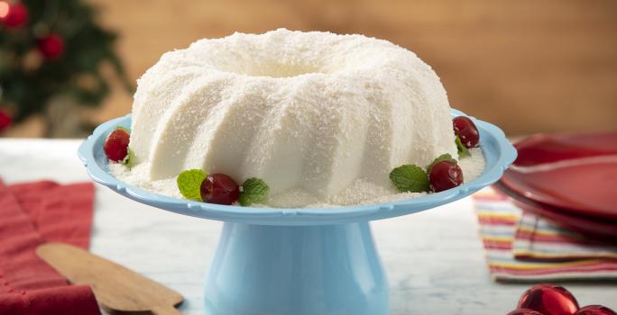 Foto da receita de Sobremesa Gelada de Coco. Observa-se uma boleira azul com a sobremesa em cima, polvilhada de coco e enfeitada com cerejas na base. Decorações de Natal enfeitam a foto, como a árvore no fundo desfocada e bolas natalinas.