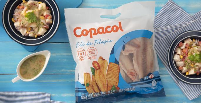 Foto da receita de Ceviche Refrescante com Filé de Tilápia Copacol, vista de cima, servido em uma tigela azul, ao lado de uma molheira branca, sobre uma bancada azul com tecidos e com um pacote do produto Copacol