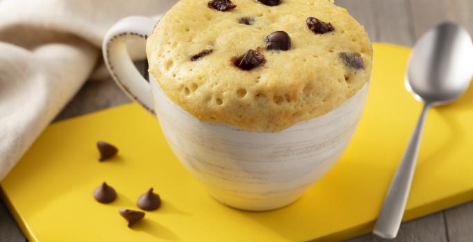 Fotografia em tons de marrom. branco e amarelo de uma bancada de madeira com uma tábua amarela, sobre ela uma xícara branca com bolo de caneca e uma colher. Ao fundo um paninho branco.