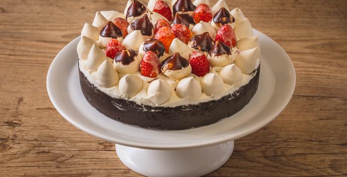 Foto aproximada de uma torta num prato branco alto. A torta tem uma base de biscoito escura e está decorada com chantilly, suspiros pequenos e morango, alguns banhados em chocolate. Tudo está numa bancada de madeira.