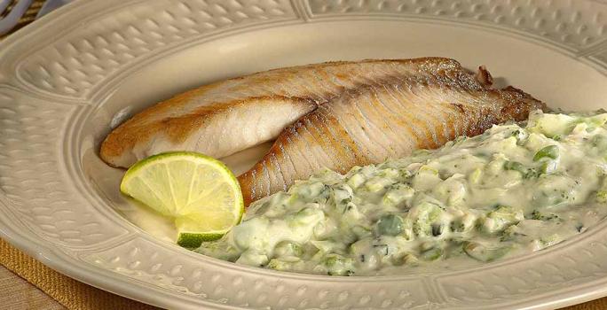 Fotografia em tons de verde em uma mesa de madeira, com um jogo americano amarelo, um prato redondo bege com o filé de peixe e o purê de brócolis dentro dele.