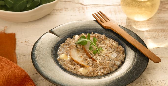 Foto da receita de risoto de pera com brie servida em um prato fundo azul com um garfo de madeira na borda e um copo de vidro com vinho branco ao fundo