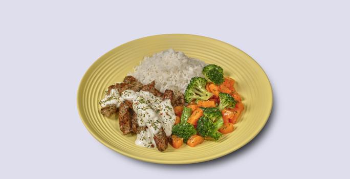 Fotografia em tons de amarelo em um fundo branco com um prato amarelo com arroz, legumes salteados e tirinhas de carne com molho de iogurte.