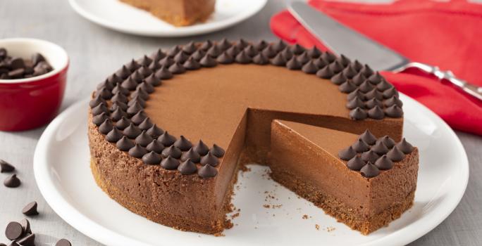 Fotografia em tons de branco e vermelho de uma bancada branca com um prato branco, sobre ele uma torta de chocolate. Ao lado um paninho vermelho com uma espátula. Ao fundo um prato branco com uma fatia de torta e um potinho com gotas de chocolate.