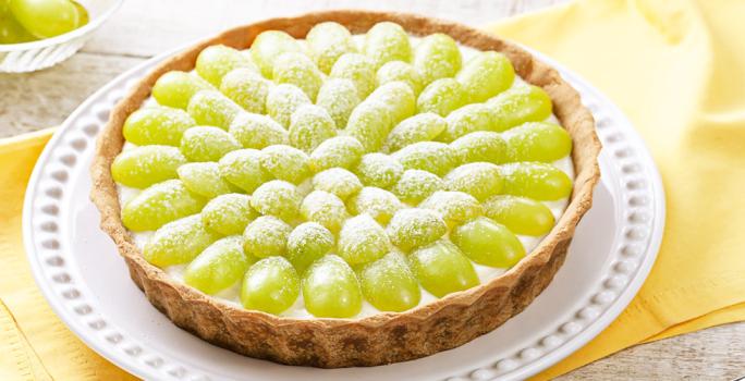 Fotografia em tons de verde e branco, prato branco ao centro contendo uma torta de uva sobre guardanapo amarelo, ao lado potinho com uvas, tudo sobre bancada de madeira com tons em branco