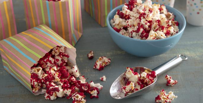 Foto da receita pipoca doce vermelha. Observa-se uma tigela azul com a pipoca e, ao lado, saquinhos estampados com a pipoca também.