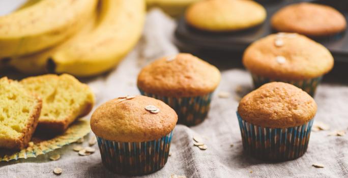 Fotografia de quatro muffins de banana e aveia dentro de uma forminha de papel colorida sobre uma toalha de mesa em tons de bege. Ao lado, um cacho de bananas, duas fatias do bolo, e ao fundo, dois muffins em uma forma preta de bolinhos.