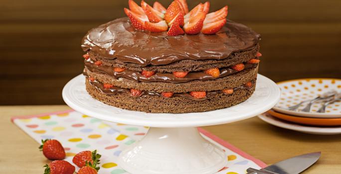 Fotografia em tons de marrom e vermelho de uma bancada de madeira, uma boleira com o bolo de chocolate e morangos. Ao lado quatro morangos para decorar, um espátula para servir e três pratos coloridos em tons de laranja com garfos.