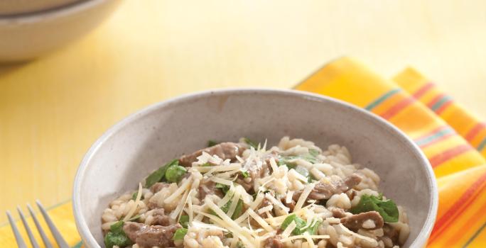 fotografia em tons de bege, cinza e amarelo de uma bancada bege vista de cima. Contém um pano amarelo com um recipiente redondo cinza com o risoto, com pedaços de carne e ao lado um garfo para servir.