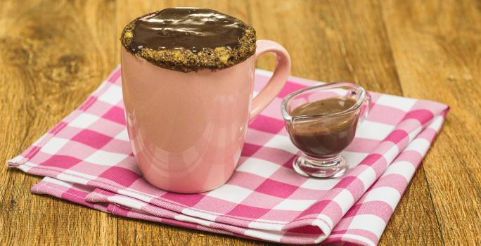 Fotografia em tons de rosa em uma bancada de madeira escura, um pano rosa xadrez, uma caneca rosa com um bolo de caneca de banana com mel, aveia e cobertura de chocolate. Ao lado, um potinho de vidro com a calda de chocolate.