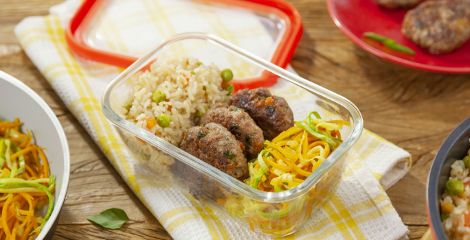 Fotografia em tons de amarelo em uma mesa de madeira com um pano de prato branco com listras amarelas, um potinho de marmita de vidro com o arroz integral, o mini hambúrguer e o macarrão de legumes