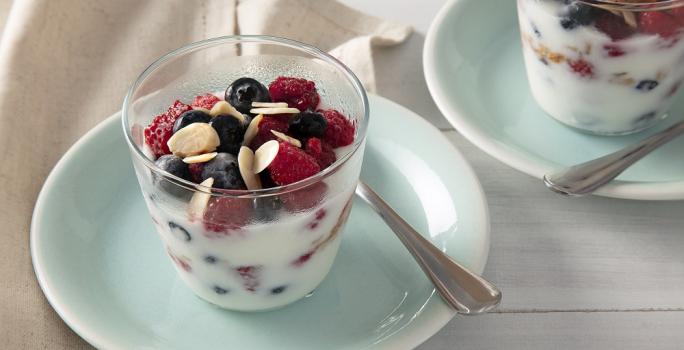 Foto em tons de azul claro da receita de parfait de iogurte com frutas vermelhas servida em dois copos altos sobre pratos pequenos azuis
