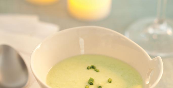 Fotografia em tons de verde em uma mesa de madeira com uma toalha verde, ao centro um potinho branco redondo fundo com o creme de abobrinha com cebolinha decorando. Ao fundo, duas velas acesas e uma taça de vidro com água.