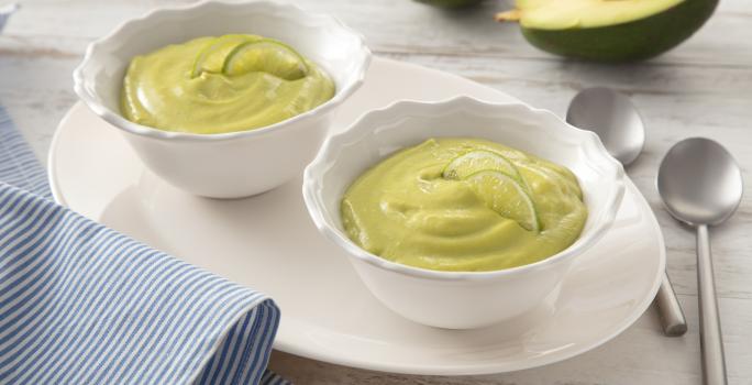Fotografia em tons de branco e verde de uma bancada, ao centro dois potinhos com creme verde de abacate. Ao fundo, um abacate cortado ao meio.